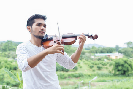 年轻的嘻哈音乐家男子拉小提琴在大自然户外生活背后的山。