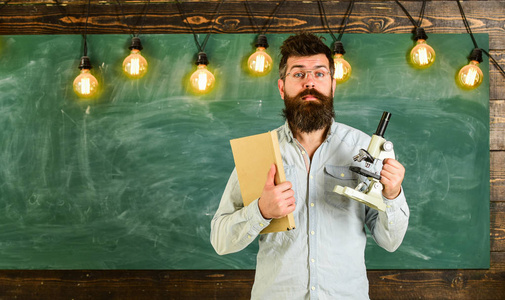 戴眼镜的老师手持书本和显微镜。在教室里惊讶的脸上留着胡子和胡子的男人。科研理念。科学家持有书和显微镜, 背景黑板