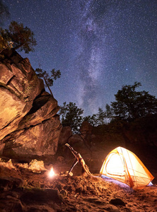 在巨大的陡峭岩层中的营地。 由篝火女子点燃，在晴朗的夜空星空背景下，在小旅游帐篷附近做瑜伽练习。 健康的生活方式概念。 阿多穆