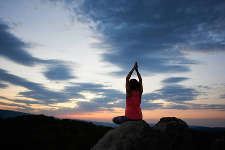背部的年轻苗条运动妇女坐在大岩石上瑜伽姿势，举起手臂在绿色森林和美丽的天空在日落背景。 冥想和健康的生活方式概念。 希德哈萨纳
