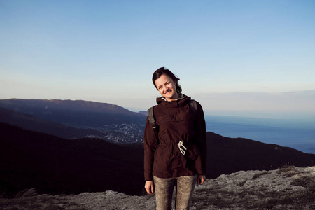 一个穿着夹克背着背包的女孩站在山顶上, 在落日的光线下俯瞰大海