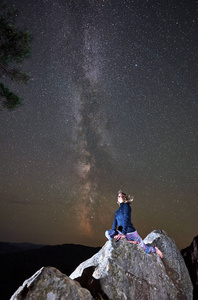 年轻苗条的旅游妇女独自坐在巨大的巨石上，夜空下星光灿烂，享受着山上的宁静。旅游旅游远足娱乐和积极生活方式的概念。