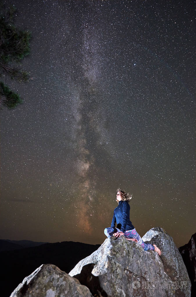 年轻苗条的旅游妇女独自坐在巨大的巨石上夜空下星光灿烂享受着山上的