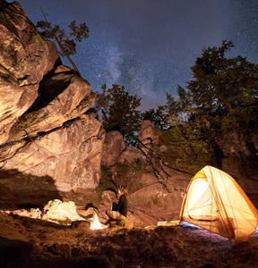 晚上在巨大的陡峭岩层中的营地。 年轻的女孩在篝火上休息，在清澈黑暗的星空背景下，在旅游帐篷前。 旅游体育和野营理念。