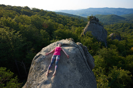 躺尸在明亮温暖的夏日,年轻女子躺在绿色森林顶部的大山岩上放松的