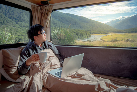 年轻的亚洲人喝着热咖啡，在露营车里的床上用笔记本电脑工作，通过窗户看到山景，数字游牧在路上旅行的概念