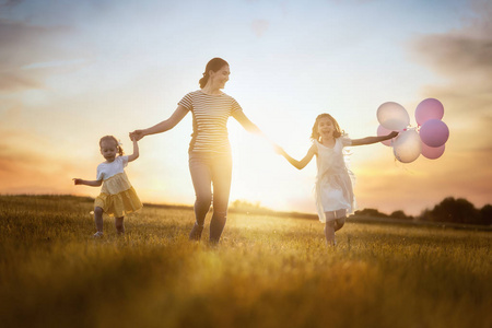 幸福的爱的家庭在夏天在大自然上玩得很开心。 年轻的母亲和两个女儿在日落的背景下在草地上欢笑和玩耍。