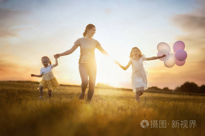 年轻的母亲和两个女儿在日落的背景下在草地上欢笑和玩耍.