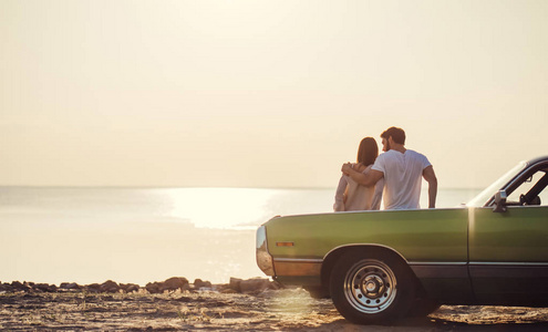 浪漫夫妇站在海滩上的绿色复古车附近。 英俊的胡须男人和迷人的年轻女人与老式的经典汽车。 爱情故事。