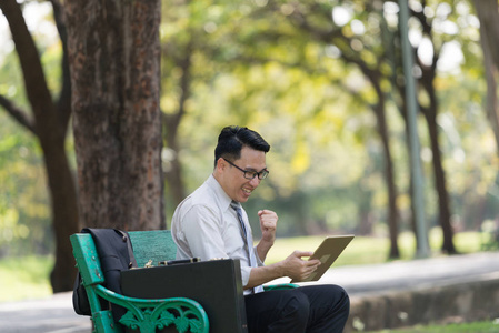年轻的商人在公园里看数字平板电脑。他对他们的成功表现出高兴的迹象.一个成功的商人概念