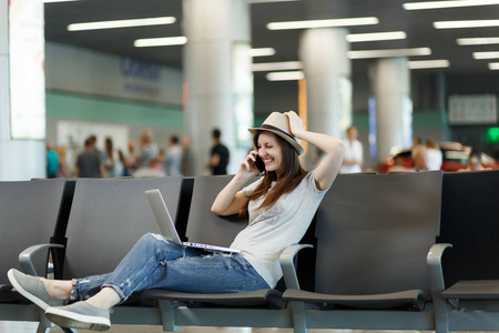 年轻的笑客旅游妇女在手提电脑上工作，在手机上打电话，朋友预订出租车酒店，在机场大厅等着。 周末出国旅行的乘客。 空中飞行概念
