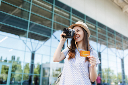 年轻微笑的旅行者旅游妇女戴着帽子，在复古老式照片相机上拍照，在国际机场持有信用卡。 周末出国旅行的乘客下车。 空中飞行概念