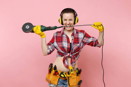 强大的能量杂工妇女在黄色手套，噪音绝缘耳机套件工具，皮带，充满仪器，持有电源锯隔离在粉红色背景。 女性从事男性工作。 翻修概念