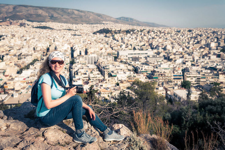 可爱的年轻女子从上面拍了一张雅典城市景观的照片，并在镜头希腊微笑。 女旅行者坐在山顶上俯瞰雅典城。 雅典的山丘是旅游景点。