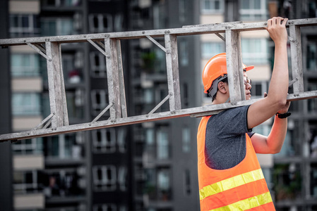 年轻的亚洲维修工人，带橙色安全帽和背心，在施工现场携带铝步梯。 土木工程建筑建设者和建筑服务理念