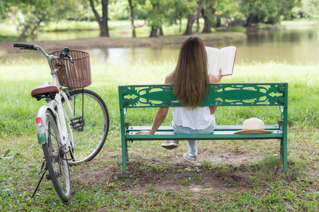 绿色阅读亚洲妇女的后肖像阅读教科书,坐在自行车和池塘附近的绿色
