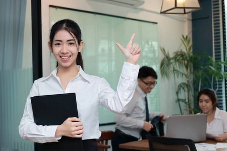 领导商业女性的概念。 开朗的年轻亚洲女商人，带着戒指的活页夹站在她的同事的办公室背景下。
