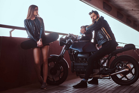 一对有吸引力的夫妇的肖像，一个穿着黑色皮夹克的野蛮留胡子的自行车手，坐在摩托车上，他年轻的黑发女朋友抱着滑板在桥下看着摄像机