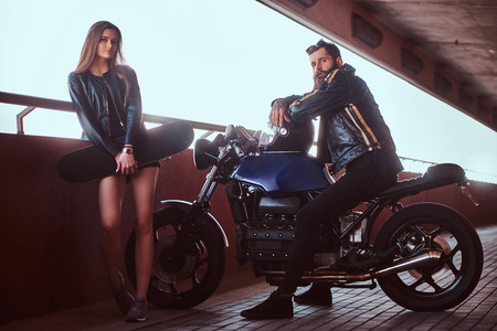 一对有吸引力的夫妇的肖像，一个穿着黑色皮夹克的野蛮留胡子的自行车手，坐在摩托车上，他年轻的黑发女朋友抱着滑板在桥下看着摄像机