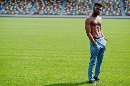 英俊的非裔美国人，肌肉发达，的躯干，穿着牛仔裤工作服，站在体育场足球场的绿草上。 时尚的黑人肖像。