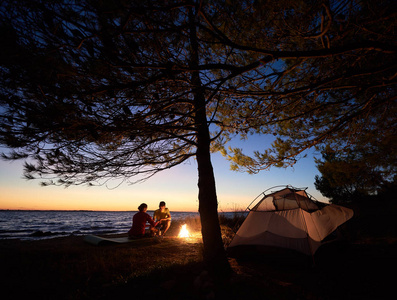 晚上在海边露营。 在树下的旅游帐篷和年轻的夫妇，男人和女人在篝火旁准备食物，在夜空和水的背景下。 旅游和冒险概念。