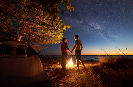 在蓝色星空下的湖边露营。 背面的年轻游客，男人和女人站在篝火上，清澈的水和城市的灯光在遥远的海岸背景。 旅游概念