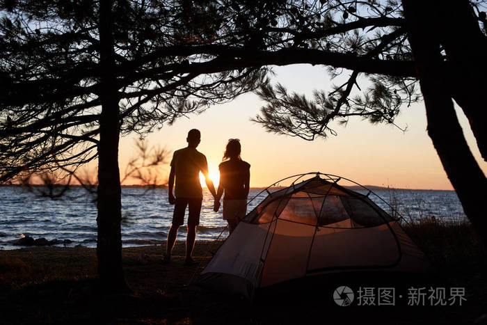 黑暗的轮廓，年轻的旅游夫妇，男人和女人站在湖岸上，双手握在小帐篷前，在明亮的黄色夕阳和晶莹的蓝色清澈的湖水背景下。