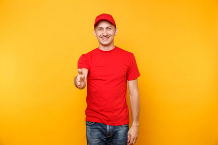 送货员穿红色制服隔离在黄色橙色背景上。 微笑的男员工戴着t帽衬衫，作为快递经销商，伸出手来打招呼。 服务概念