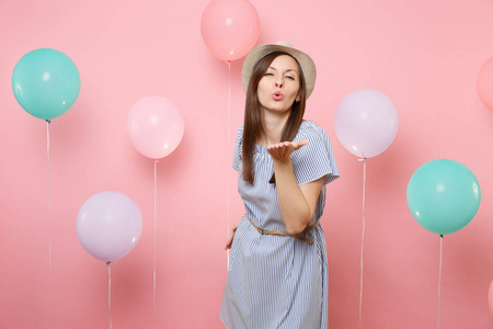 迷人的年轻女子的肖像，戴着稻草夏帽和蓝色连衣裙，用五颜六色的气球在粉红色的背景上吹着嘴唇，送着空气亲吻。 例如生日聚会真诚的情