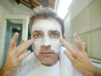 近脸肖像年轻英俊迷人的白种人在家看着自己在浴室镜子上应用白色面罩在男性美容和面膜卫生概念。