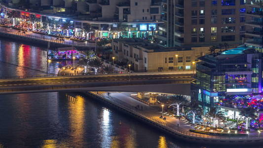 迪拜码头天际线上的运河和长廊在夜间时间推移。 具有照明和照明的住宅塔。 在摩天大楼附近的桥上漂浮的游艇和船只