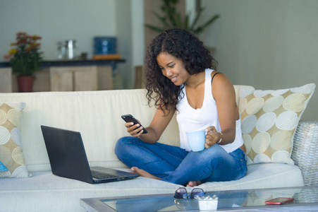 生活方式肖像年轻，快乐，美丽的黑人非洲美国妇女使用互联网短信在手机上工作时，笔记本电脑在家客厅沙发沙发放松。