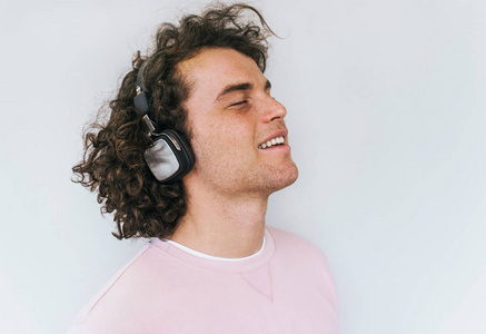 侧视肖像英俊微笑的年轻男性模特卷发和雀斑穿着粉红色的衣服，听音乐的耳机隔离在白色的工作室墙上。 广告复制空间