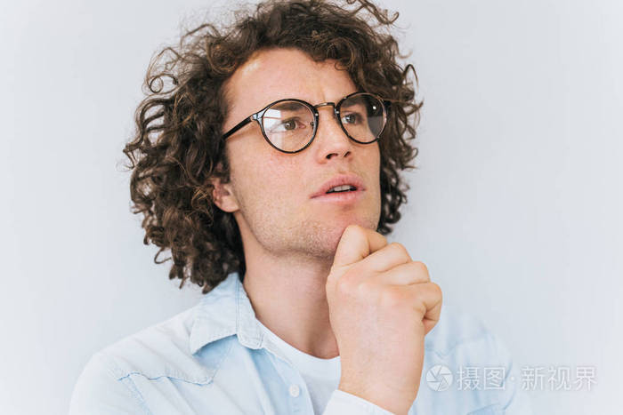 英俊的沉思男性卷发,穿蓝色衬衫和时髦的眼镜抬头,认为他的下巴在白色