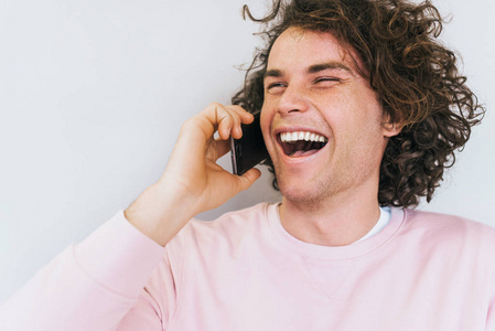 快乐开朗的卷发男性特写肖像通过手机与朋友沟通。 英俊的男人通过智能手机微笑和大笑。 人的技术概念。