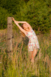 美丽的年轻女子站在田野附近的一个混凝土柱, 绿色的草和花朵。户外享受大自然。健康微笑的女孩站在高高的草丛中