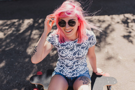 有趣的顶部景色运动美丽的微笑妇女与吹粉色头发，戴黑色太阳镜，纹身，坐在她的长板上等待她的朋友在公园。生活方式和运动理念..