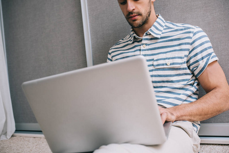 裁剪镜头，开朗的年轻欧洲男人，穿着时尚的衬衫，坐在地板上，在家里，笔记本电脑在他的腿上，有视频呼叫商业创业。 人和生活方式的概念