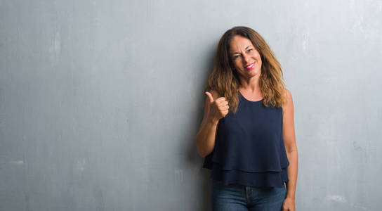 中年西班牙裔妇女站在灰色的墙壁上，用手做着快乐的竖起大拇指的手势。 赞许的表情看着相机，显示出成功。
