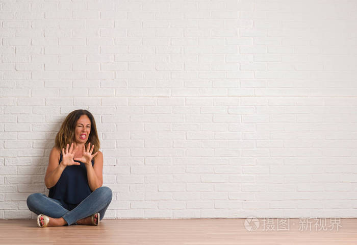 中年西班牙裔妇女坐在白色砖墙上的地板上，厌恶的表情，不高兴和恐惧的做厌恶的脸，因为厌恶的反应。 举起双手。 令人讨厌的概念。