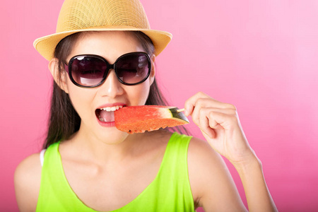 一个快乐迷人的女人的肖像，穿着夏天的绿色衣服，戴着帽子和太阳镜，在粉红色的背景下，在棍子上咬着新鲜的西瓜。 夏季氛围概念。