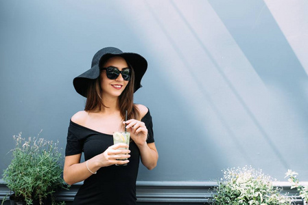 年轻漂亮的女孩戴着黑色帽子坐在街上的咖啡馆里喝着冷却的饮料。