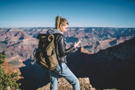 微笑的美国女游客在智能手机上阅读信息，对山区良好的数据连接感到满意，带着背包的女人，看着手中的手机，站在野外徒步旅行的悬崖上