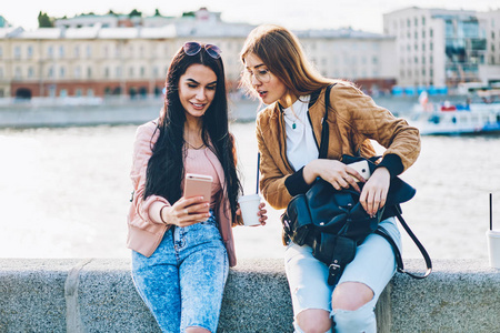 两个开朗的好朋友，带着时尚的黑色背包和美味的咖啡，通过安装在城市宣传背景上的现代电话上的新照片应用程序制作自拍照片