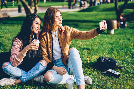 快乐的最好的朋友摆姿势在智能手机相机上自拍花时间在公园里。穿着时髦服装的快乐时髦女孩通过移动设备拍照上传到社交网络上