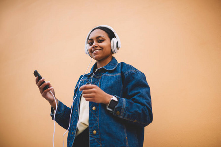 快乐的美国黑人女孩穿着牛仔裤夹克和耳机，享受最喜欢的歌曲，同时站在背景上与复制空间。 微笑女性黑色模特听音乐通过应用手机