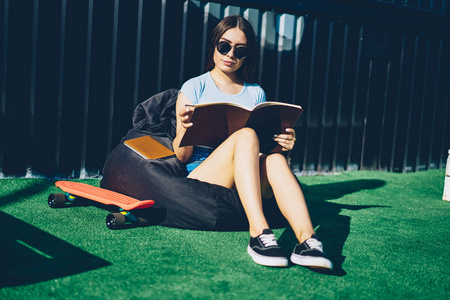 戴着时尚太阳镜的沉思的年轻女子正在阅读学习课本，舒适地撅着嘴在草坪上享受暑假。戴着黑色眼镜的时尚学生在书上消磨时间