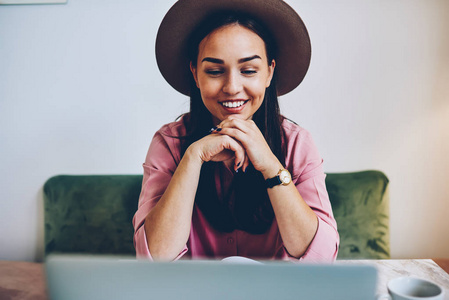 戴着时尚帽子的微笑年轻女性在空闲时间使用笔记本电脑观看在线电影，积极的女性对自由职业者在咖啡厅内阅读上网本上的新闻感到满意