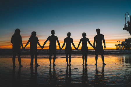剪影的年轻朋友牵着手一起欣赏美丽的风景日落在海洋海岸，一群人站在海滩上黄昏。 幸福成功友谊社区观念