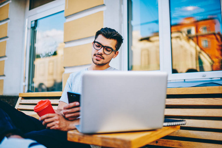 一位戴眼镜的男性博客作者在智能手机上阅读通知，同时在城市环境中远程工作，在连接到免费4g互联网的现代笔记本电脑上，坐在木制长凳上
